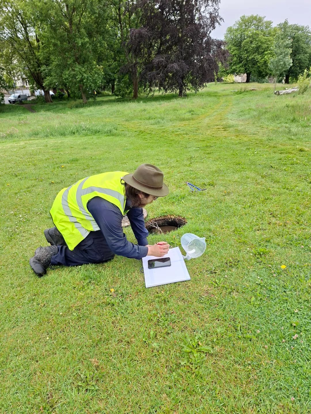 A person in a fedora hat and safety vest kneels on grass, writing in a notebook next to a small hole in the ground.